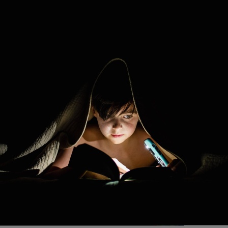 Ein Kind liegt im Bett und liest unter der Decke ein Buch bei Nacht