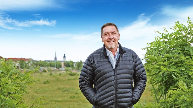 Der Moderator "Böttcher" steht in Leipzig auf einer grünen Brachfläche.