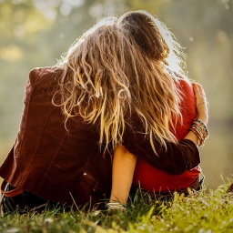 Zwei Frauen sitzen am See, von der die eine ihren Arm um die Schulter der anderen legt.