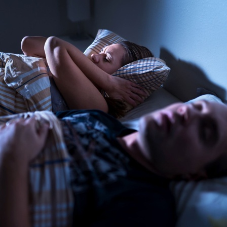 Eine Frau liegt neben ihrem schnarchenden Mann im Bett und hält sich die Ohren zu. 