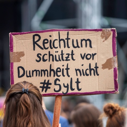 Bei einer Demonstration hält eine Teilnehmerin ein Schild mit der Aufschrift "Reichtum schützt vor Dummheit nicht #Sylt" hoch.