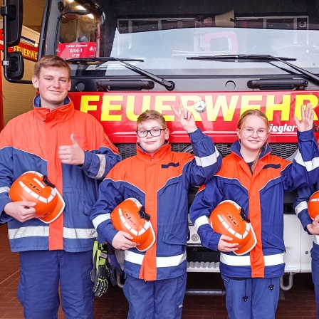 Ein Jugendlicher und drei Kinder stehen vor einem Feuerwehrauto und winken.