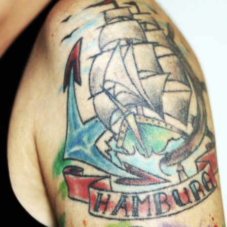 Farbige Tätowierung auf einem Oberarm mit einem Segelschiff und dem Schriftzug Hamburg.