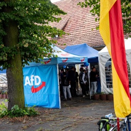 Unterstützer der AfD stehen bei einer Wahlparty der AfD in einem blauen Zeltpavillon mit dem Logo der Partei, davor weht eine Deutschlandflagge.