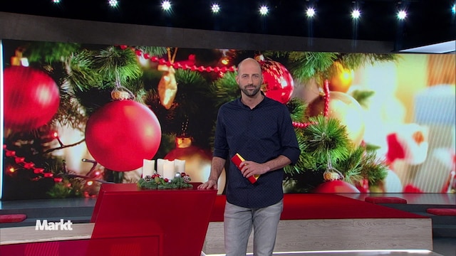 Auf dem Bild sieht man den Moderator Simon Pützstück im Markt Studio vor einem Bild von einem Geschmückten Weihnachtsbaum