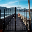 Blick auf einen Steg, der auf einen sonnenbeschienenen See führt. Davor ein verschlossenes schmiedeeisernes Tor mit einem Schild, auf dem steht "Zutritt für Unbefugte verboten".