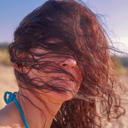 Eine Frau steht bei windigem Wetter an einem Strand. Ihre Haare bedecken ihr Gesicht.