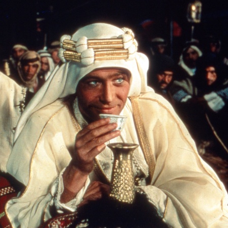 Moviestill von Lawrence von Arabien. Lawrence (Peter O&#039;Toole) sitzt im arabischen Outfit vorne im Bild und trinkt Tee.
