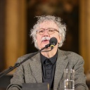 Der Schriftsteller Karl-Markus Gauß mit grauem, lockigen Haar und Brille spricht in ein Mikrofon. Vor ihm steh ein Glas Wasser.