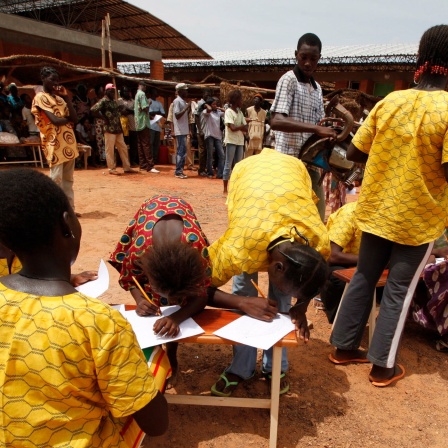 Das Operndorf Afrika zeigt Kinder in Burkina Faso, die schreiben. Im Hintergund sind Menschen versammelt. 