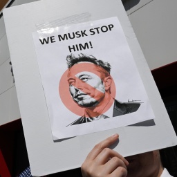 Auf einer Demonstration gegen Elon Musk / Tesla in den USA hält ein Protestant ein Schild mit der Aufschrift "WE MUSK STOP HIM!" hoch