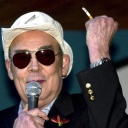 Der Hippie Chronist Hunter S. Thompson trägt Sonnenbrille und Hut, in der rechten Hand ein Mikrofo, in der linken eine Zigarette.