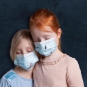 Zwei Mädchen mit geschlossenen Augen die Schutzmasken tragen, auf denen ein Smiley-Gesicht aufgemalt ist.