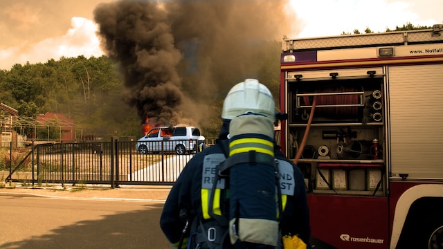 Ein Bulli brennt auf einem Grundstück, vor dem Eingangstor steht eine Einsatzkraft neben einem Löschfahrzeug