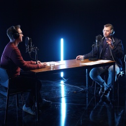 Zwei junge Männer sitzen in einem Studio an einem Holztisch und sprechen in Mikrofone, die auf dem Tisch stehen.