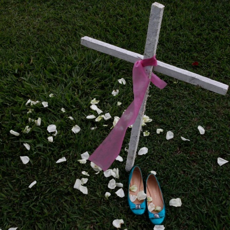 Ein weißes Holzkreuz mit einem rosa Band, daneben steht ein Paar blaue Damenschuhe.
