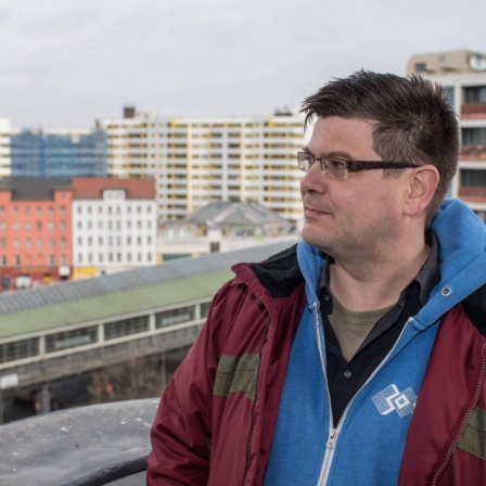 Andrej Holm steht auf einer Terrasse in Kreuzberg mit dem Wohnkomplex Zentrum Kreuzberg in seinem Rücken. 