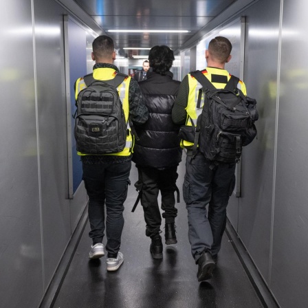 Zwei Beamte in Warnwesten eskortieren einen Mann durch einen Flugzeugtunnel.
