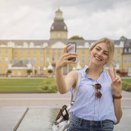 Junge Frau macht vor dem Schloss in Karlsruhe ein Selfie: Fotos von Gegenständen mit dem Handy werden uns so gezeigt, wie wir sie selbst sehen. Aber ein Selfie ist immer spiegelverkehrt. Wie kommt das?