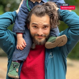 Ein Mann trägt ein Kind auf den Schultern und schaut in die Kamera.
