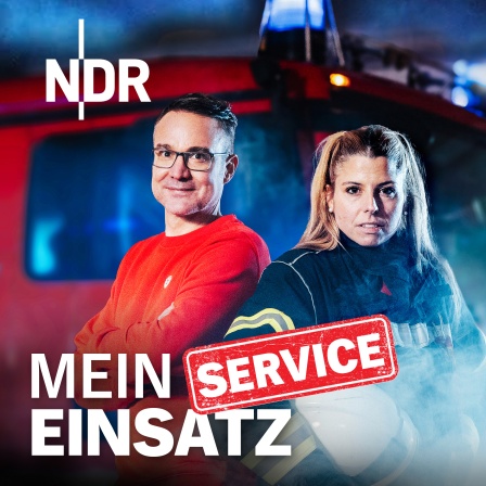 Ein Mann in rotem Pullover und eine Frau in Feuerwehruniform mit Helm stehen vor einem roten Feuerwehrauto mit Blaulicht und Rauch im Hintergrund.
