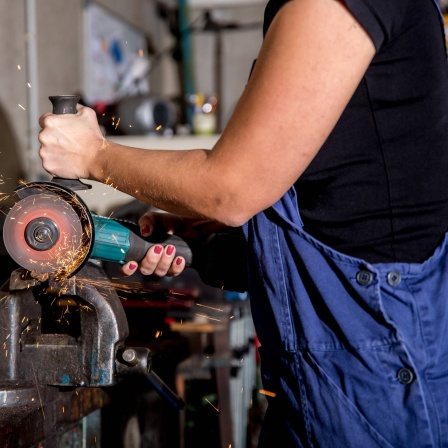Eine Frau arbeitet im Beruf als Metallbauerin in einem Betrieb des Metallbau. Mit dem Winkelschleifer schneidet Sie ein Stück Metall auf Mass.