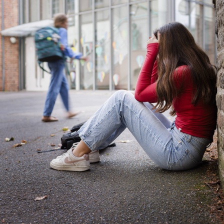 Junges Mädchen sitzt zusammengekauert an einer Mauer, mit den Händen vor dem Gesicht.