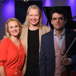 Eine blonde Frau steht neben einer anderen größeren Frau und einem Mann, der eine Geige in der Hand hält.