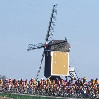 Vor einer Windmühle fahren viele Radfahrer vorbei.