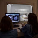 Ärzte sitzen an einem Bildschirm und werten Bilder einer Kernspintomographie aus. Im Hintergrund liegt ein Patient auf dem Tisch beim MRT.