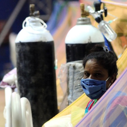 Eine Patientin sitzt auf einem Bett, sie trägt einen Mundnasenschutz. Hinter ihr ist eine Sauerstoff-Flasche zu sehen.