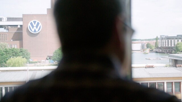 Ein unkenntlich gemachter Mann steht mit dem Rücken zur Kamera vor einem Fenster mit Blick auf den VW-Firmensitz