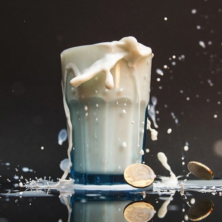 Ein Glas mit Milch, die über den Rand hinausläuft und einige Euro Geldstücke liegen auf dem Boden