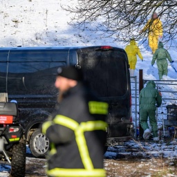 Personen in Schutzkleidung gehen über eine Weide, während Feuerwehr und Polizei das Gelände weiträumig abgesperrt haben.