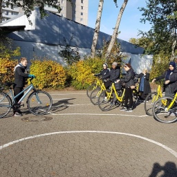 Zu sehen ist ein Schulhof mit weißen Verkehrslinien, vor einer Gruppe Frauen mit Rädern steht eine Trainerin und erklärt.