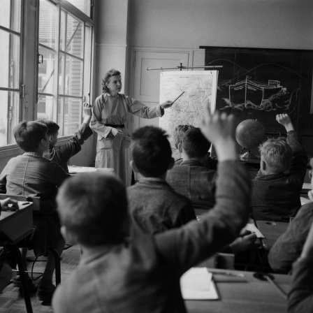 Schwarz-weiß Bild: Schülerinnen und Schüler in einer Klasse, manche strecken den Arm. Die Lehrerin erklärt etwas mit einem Zeigstock an der Tafel.