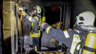 Zwei Feuerwehrmänner löschen in einem schwarzen Raum ein Feuer.