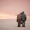 Zwei Personen in Schutzanzügen stehen zusammen in sandiger Landschaft, Gasmasken in den Händen.