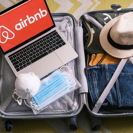 Gepackter Koffer mit Mundschutz und Laptop mit Airbnb-Logo