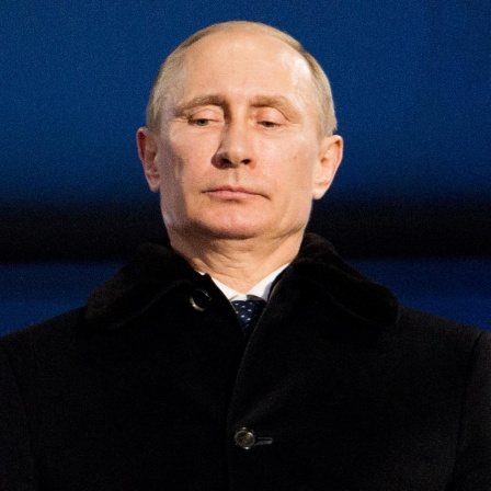Portrait von Putin, der einen dunklen Mantel trägt, vor einer blauen Wand steht und nach unten schaut.