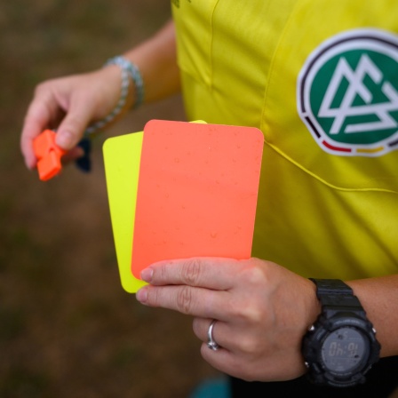 Eine Schiedsrichterin, steht mit roter und gelber Karte auf einem Rasen und hält in der anderen Hand eine Trillerpfeife.