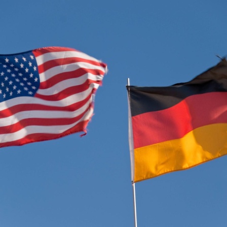 Eine US-amerikanische und ein deutsche Flagge flattern im Wind, dahinter blauer Himmel