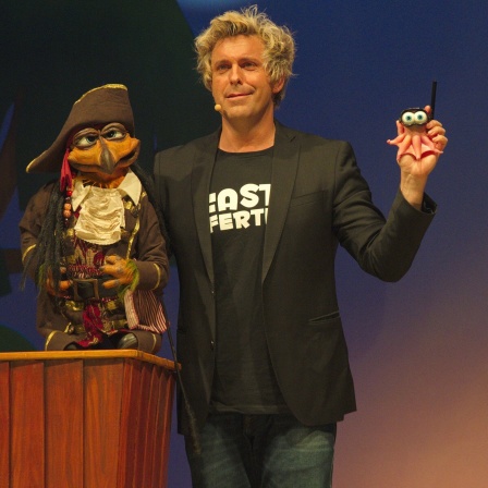 Ein Bauchredner steht auf einer Bühne und hält zwei Puppen in seinen Händen.