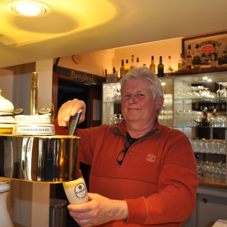 Ein Mann steht hinter dem Tresen eines Landgasthofs und zapft ein Bier.