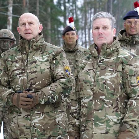Keir Starmer und John Healey stehen mit vier Soldaten des britischen Militärs in militärischen Uniformen vor einem Wald.