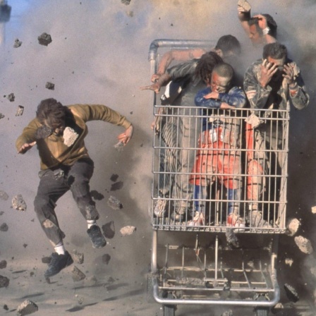 Stuntman fahren in einem vollbesetzten Einkaufswagen, während im Hintergrund etwas explodiert.