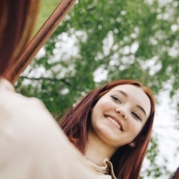 Eine junge Frau hält einen großen Spiegel in der Hand und sieht sich darin selbst. Sie lächelt.