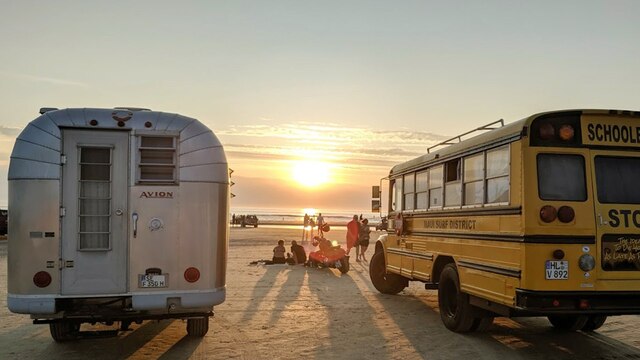 Der Schulbus-Camper und ein klassischer, silberner US-Camper stehen an einem Strand. Die Sonne geht unter.