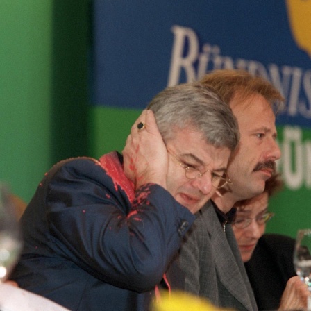 Joschka Fischer hält mit schmerzverzerrtem Gesicht seine Hand ans Ohr. Gesicht und Jackett sind mit roter Farbe beschmiert.