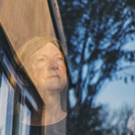 Eine ältere Frau steht am Fenster und blickt in die Ferne, in der Fenterscheibe spiegelnsich Himmel und Bäume.